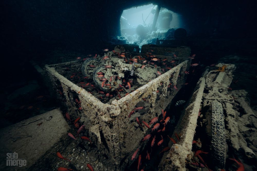 Underwater wreck covered in rust and coral formations viewed through a dark cave opening in dramatic lighting.