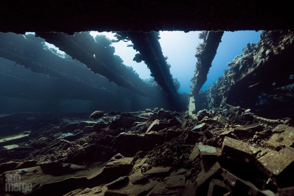 Sunlight streams through clear blue water in an underwater cave opening with rugged rock formations.