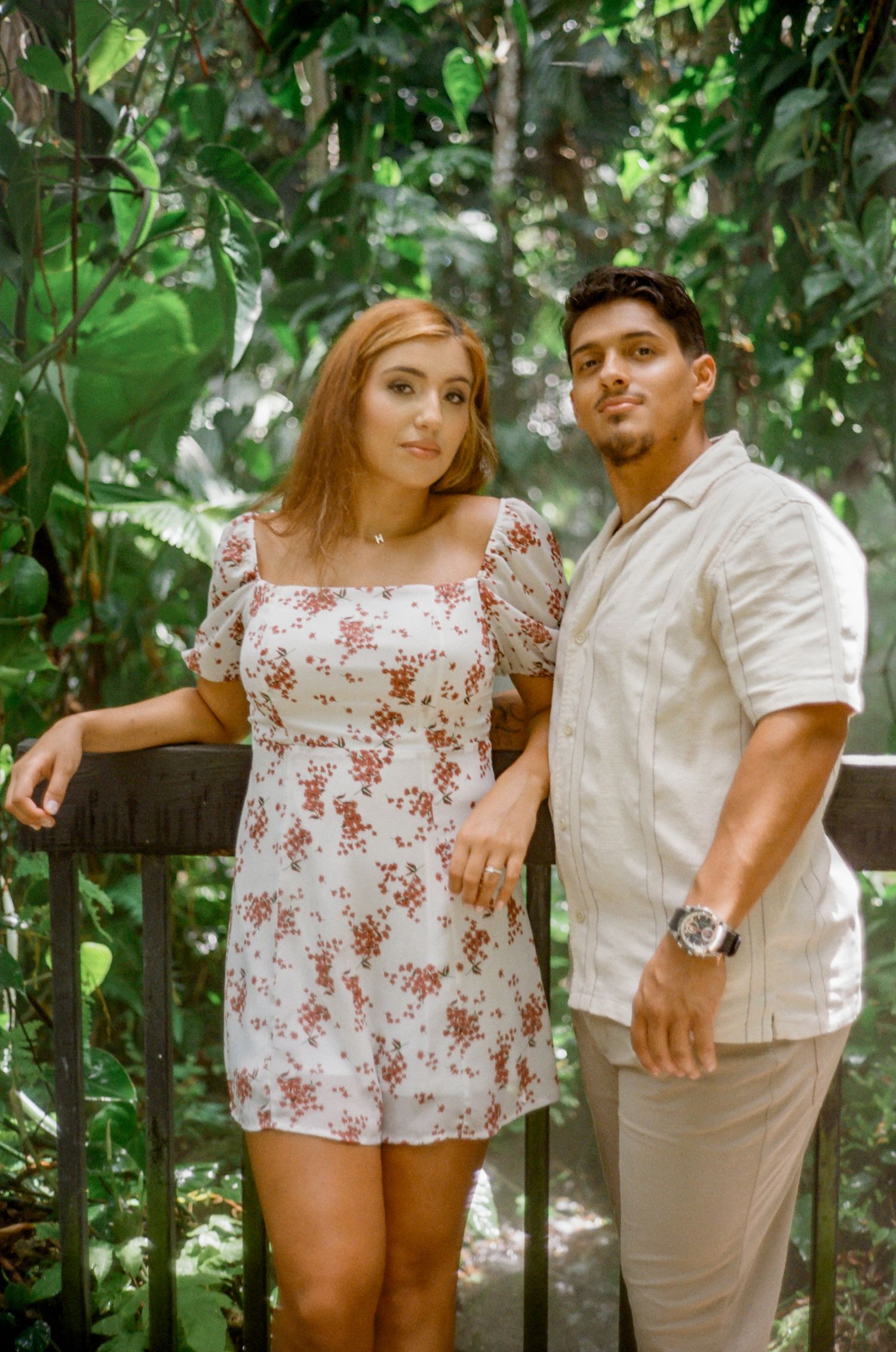 Couple poses together outdoors against green foliage backdrop in summer casual attire with floral and white clothing.