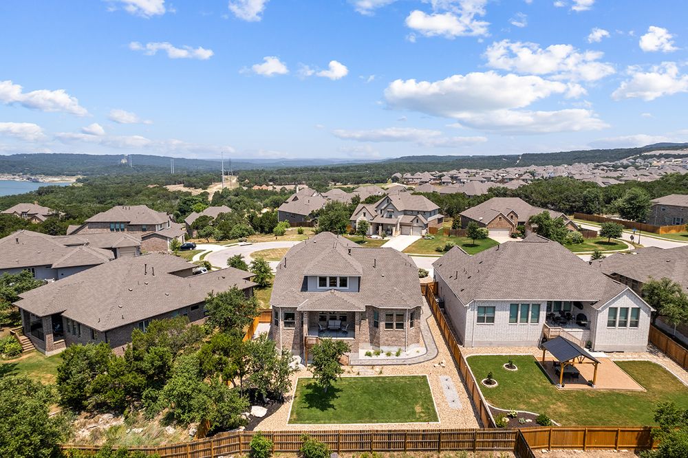 Aerial panoramic view of modern suburban homes with tan roofs and green lawns under a bright blue sky with white clouds.