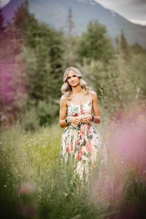 High school senior girl in a floral dress stands in a dreamy meadow with wildflowers and soft bokeh effects.