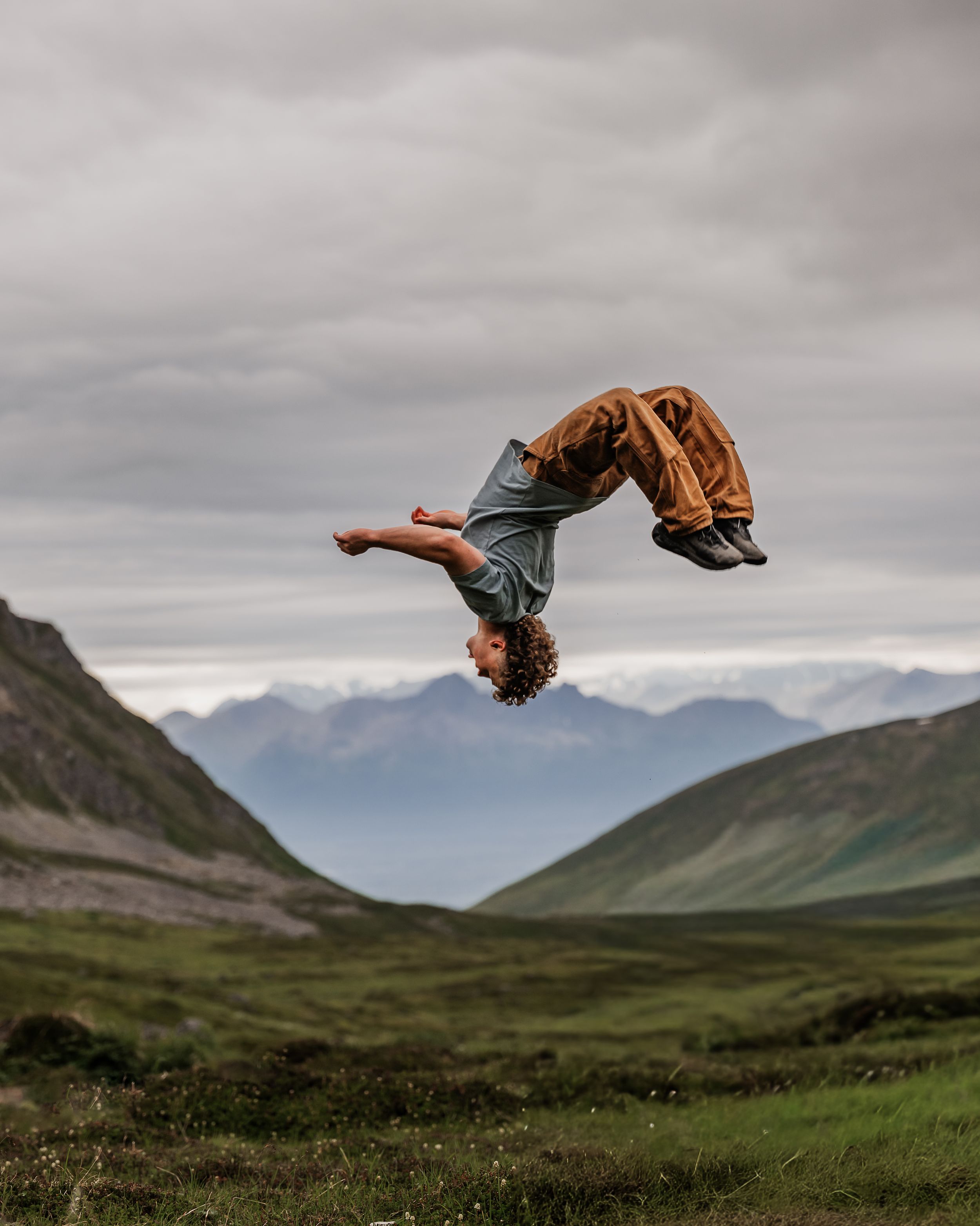 High school senior boy gymnast doing a backflip against a dramatic mountain landscape with cloudy skies.