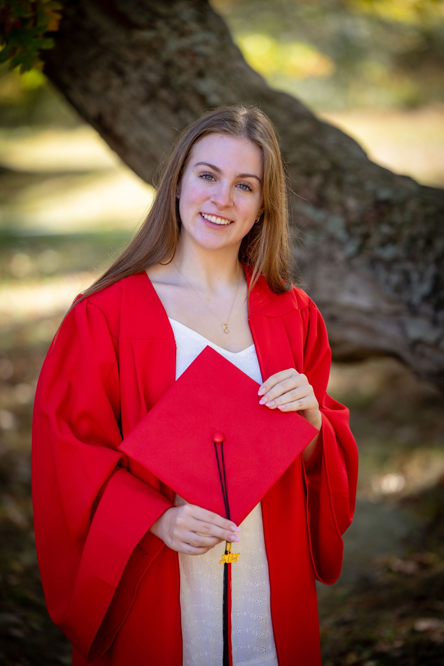 A graduate in bright red graduation gown holds their cap while smiling outdoors near a tree trunk.