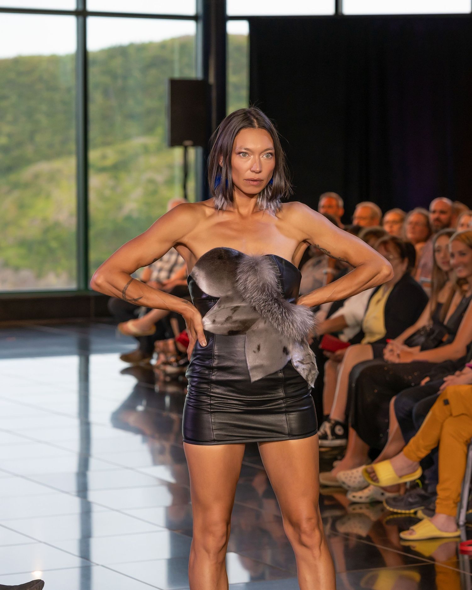 Model walks runway in strapless gray silk dress during fashion show with mountain backdrop visible through windows.