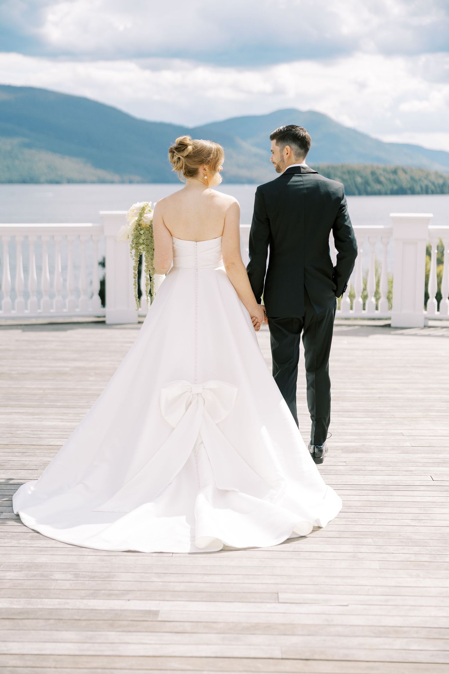 A bride in a flowing white gown and groom in black suit stand together on a deck overlooking a scenic mountain lake.
