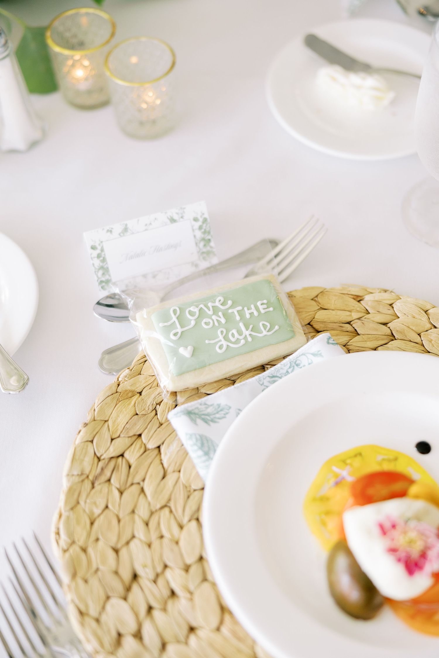 Place setting with woven placemat, white dinnerware, and soft mint green name card on elegant white tablecloth.