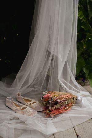 Delicate wedding shoes and bouquet styled with flowing white tulle veil on light surface.