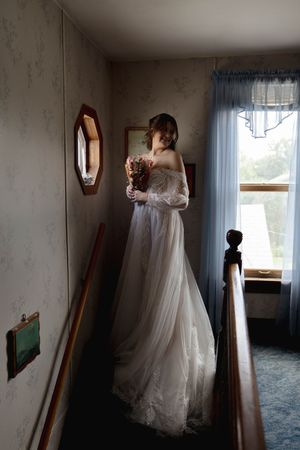 Wedding dress hangs by window in natural light with mirror reflection.