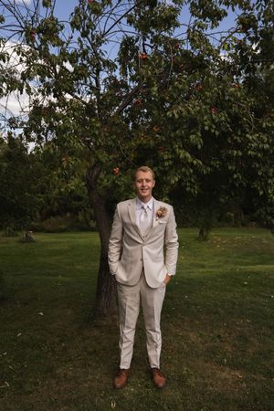 Full length portrait of someone in a cream suit standing in an orchard.