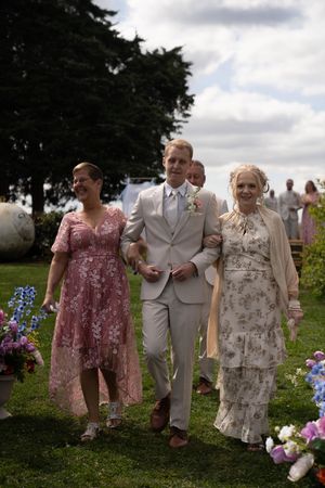 Wedding guests in floral dresses and light suits walk together on a grassy hill with purple flowers.