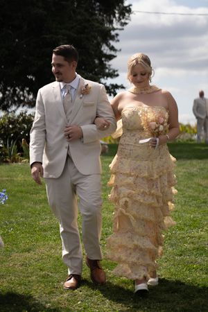 Wedding party members in beige suits and cream dresses walk together on a grassy hillside.