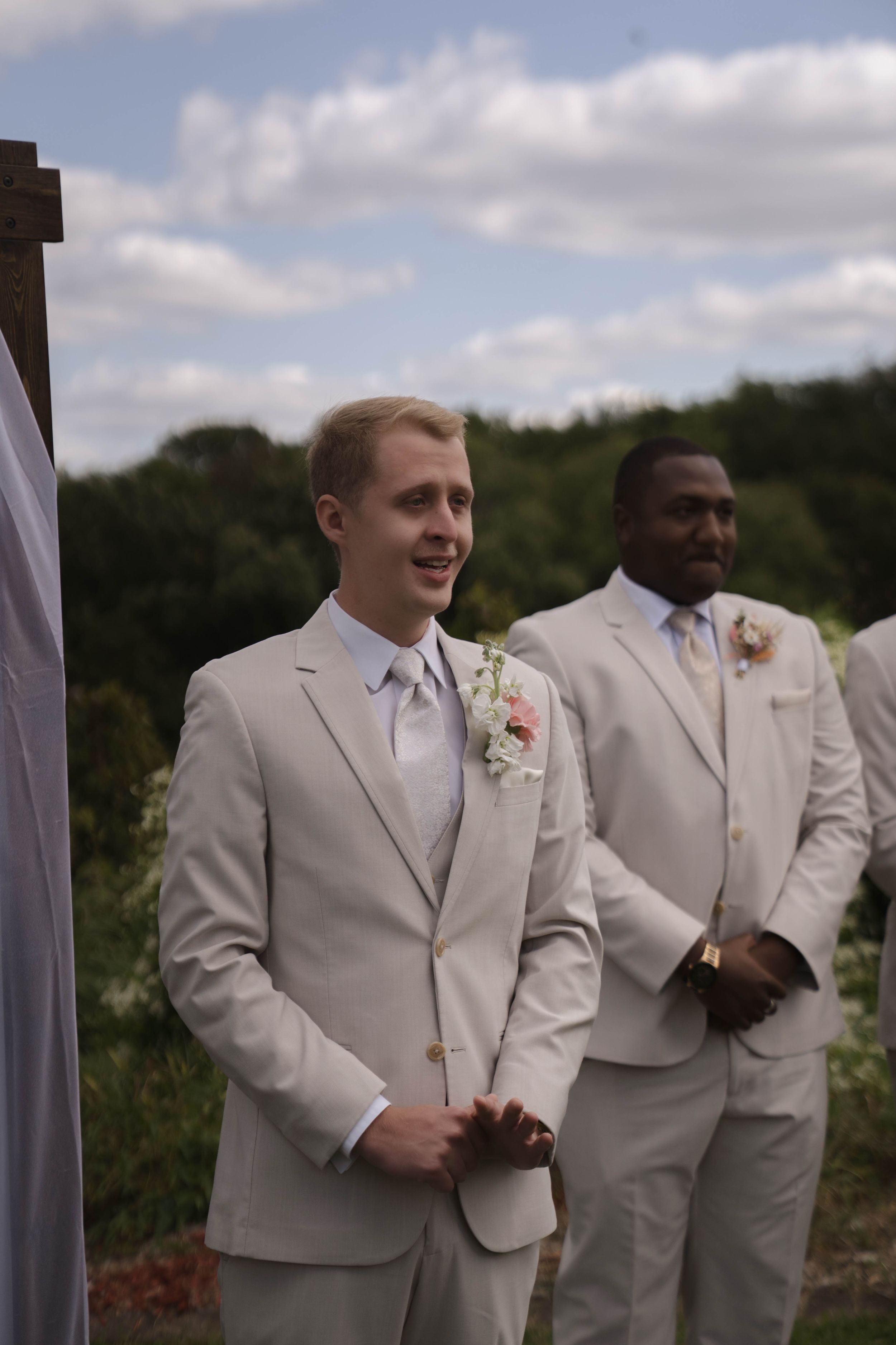 Groomsmen in light colored suits stand together during an outdoor wedding ceremony.