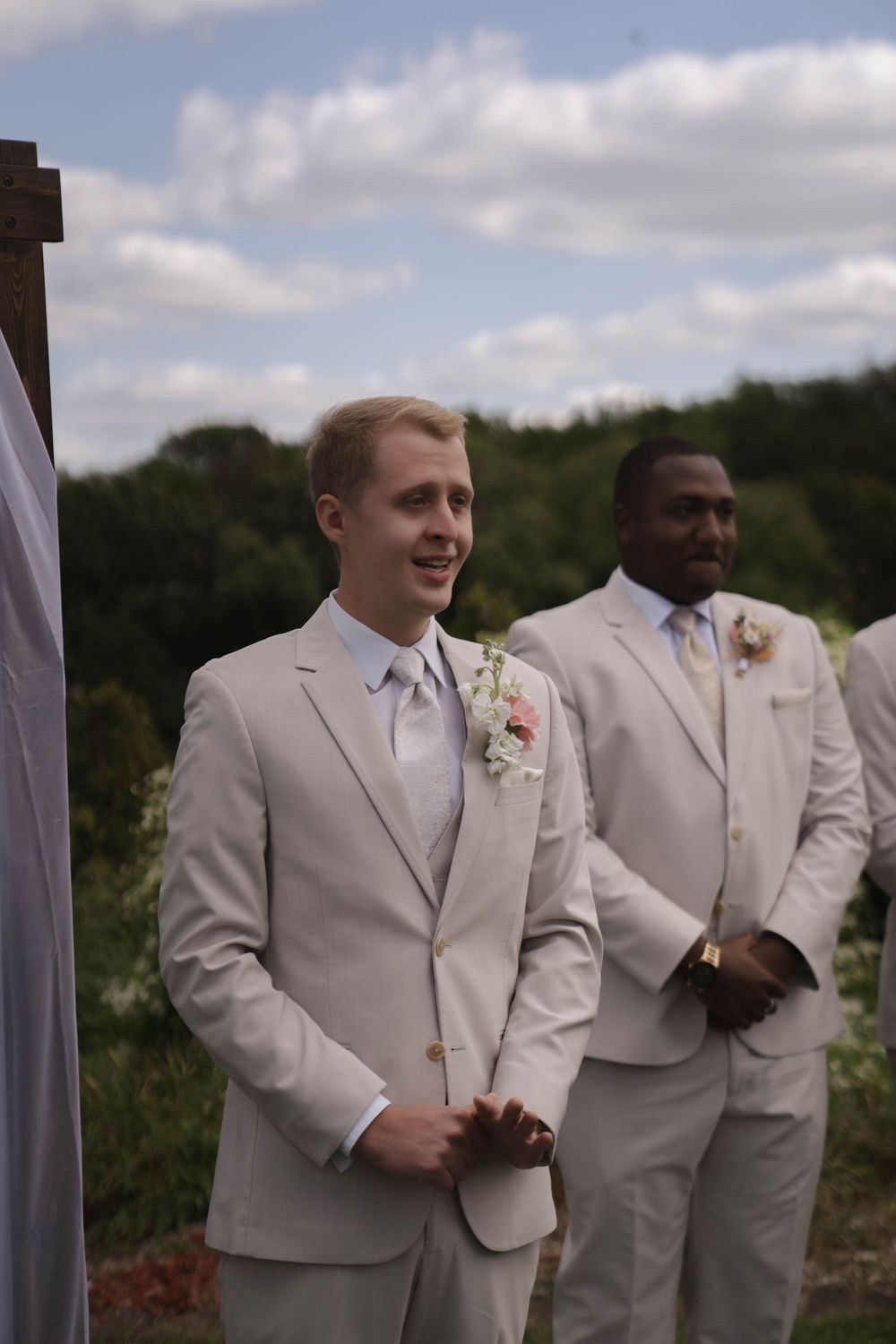 Groomsmen in light colored suits stand together during an outdoor wedding ceremony.