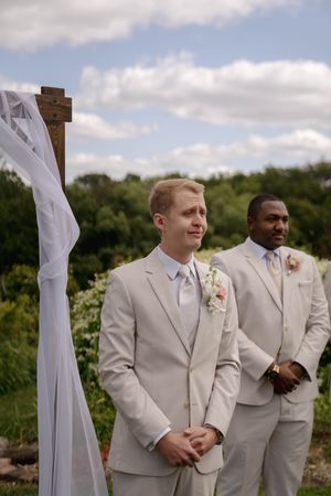 Groomsmen in light beige suits stand together during an outdoor wedding ceremony.