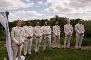 Groomsmen in light colored suits stand in a line during an outdoor wedding ceremony.