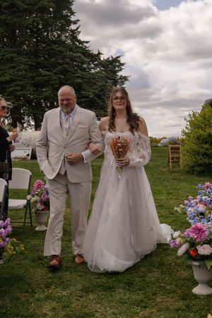Wedding party members walk together on grass with floral decorations and white chairs.