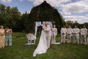 Wedding party in light blue and beige attire poses together on a grassy hill under cloudy skies.