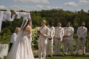 Wedding party in white attire poses outdoors in a garden setting under blue skies.