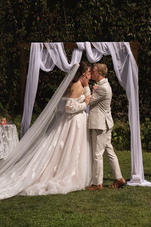 Romantic wedding scene with white draped arch and couple sharing an intimate moment outdoors.
