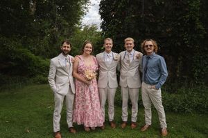 Wedding party poses casually in white suits and pink dress on garden lawn.