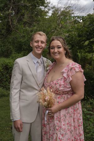 A couple poses outdoors wearing a light suit and pink floral dress during a wedding celebration.