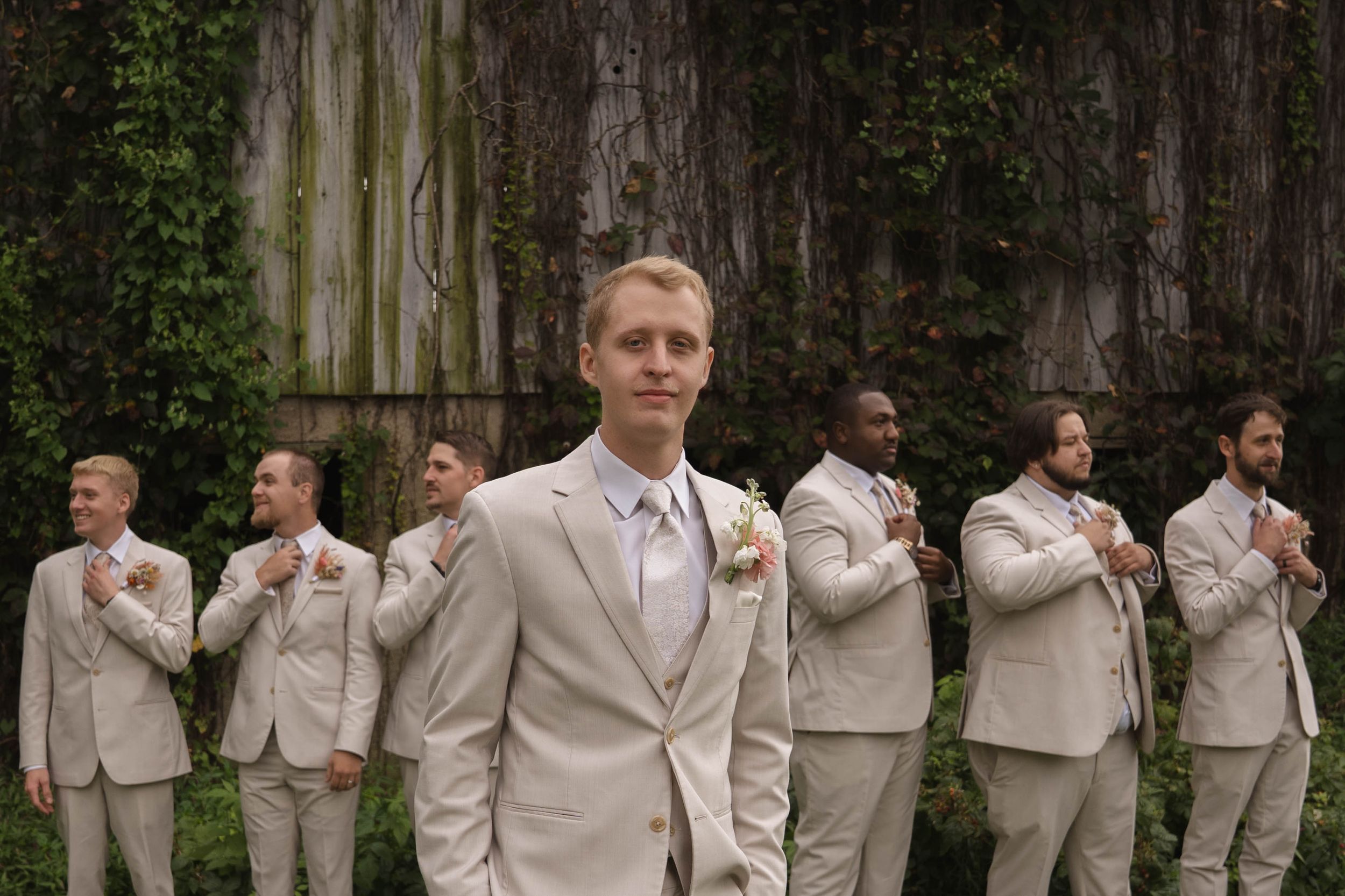 Groomsmen in beige suits pose together against rustic wall backdrop.