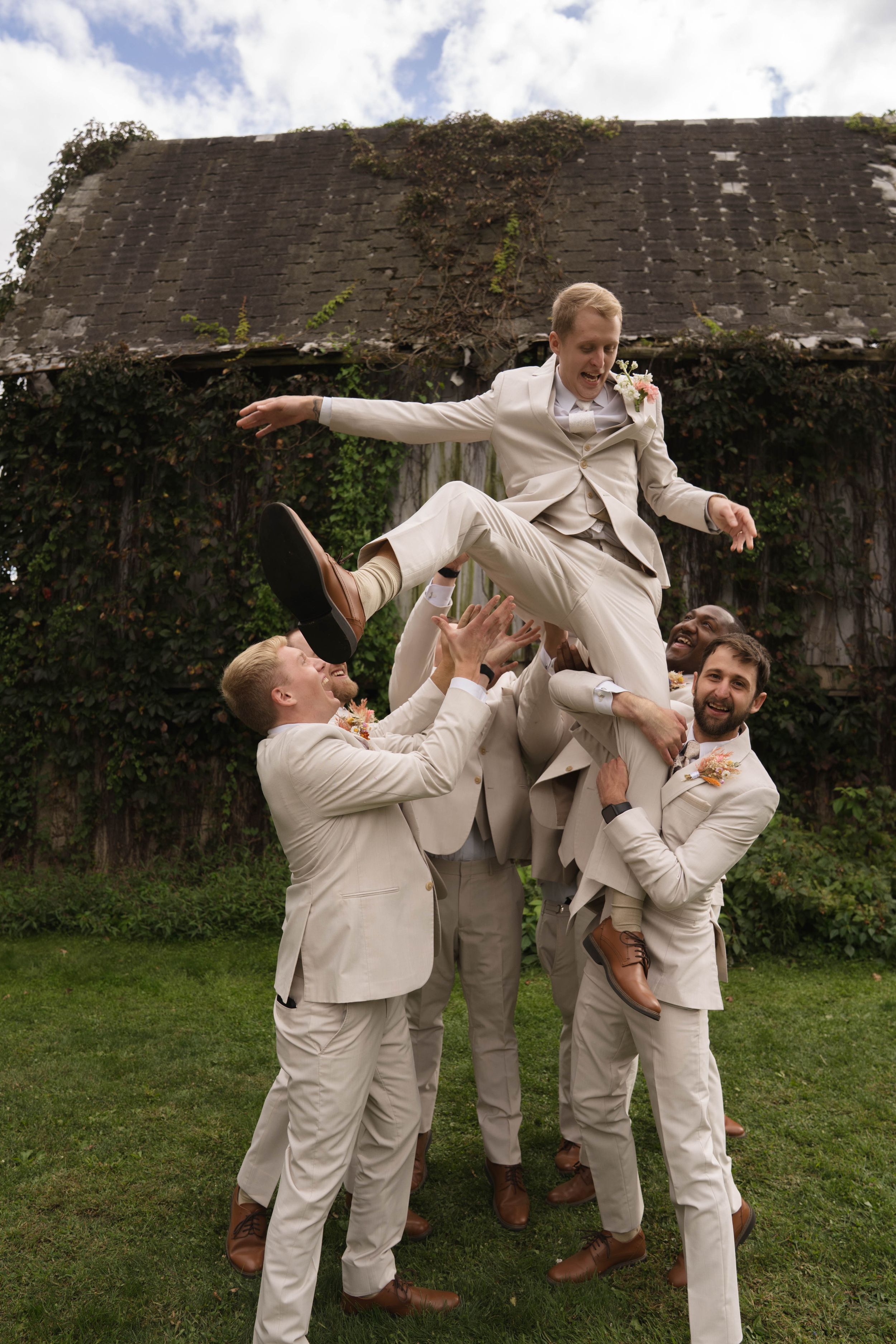 Groomsmen in beige suits perform playful lifting poses on grassy lawn.