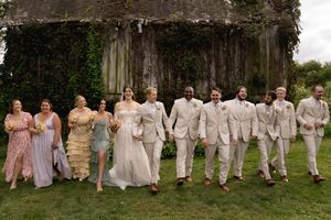Wedding party members walk together in a line across grassy lawn wearing coordinated beige and pink attire.