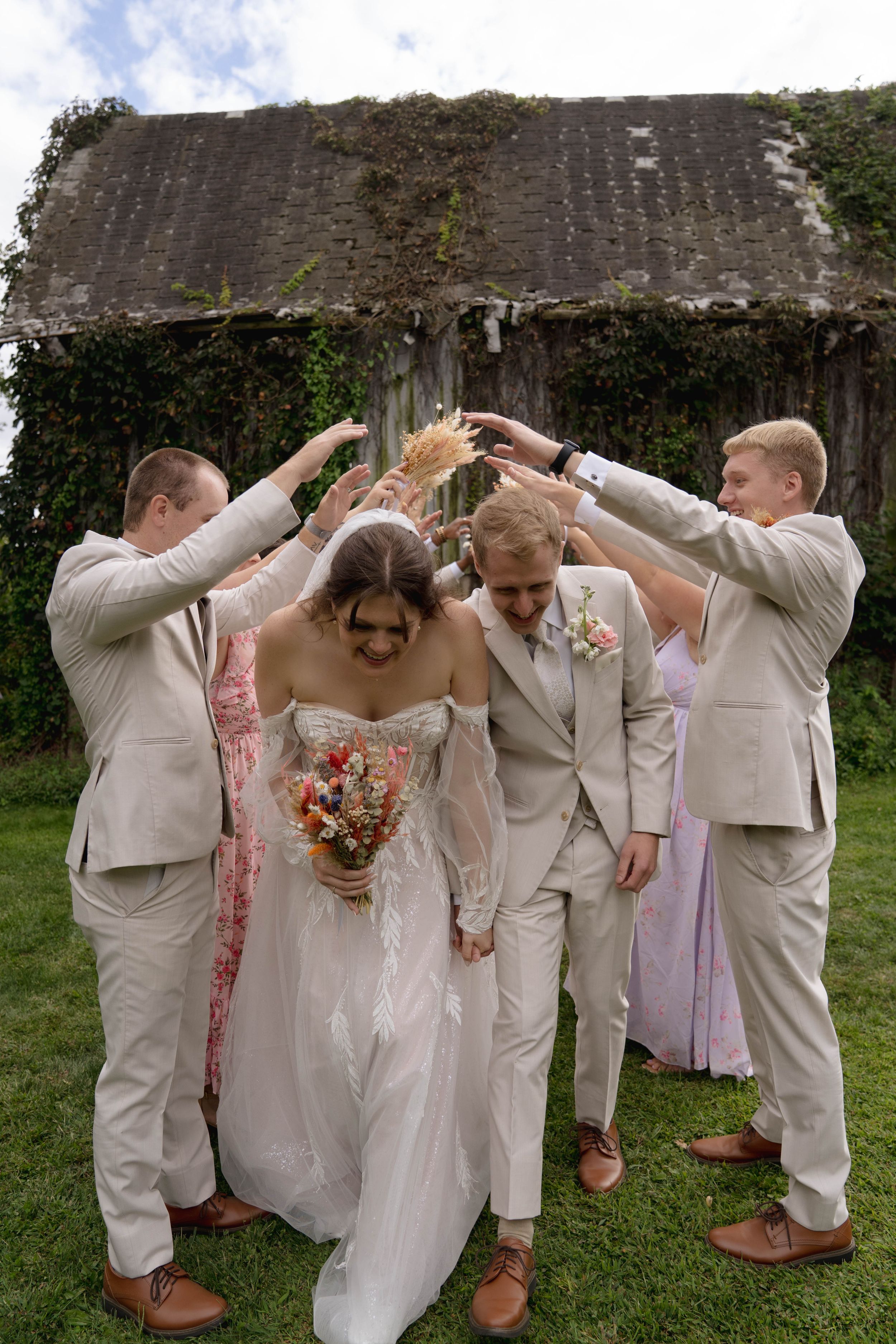 Wedding party in beige suits and floral dresses pose playfully covering their eyes in an outdoor garden setting.