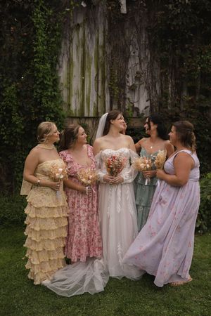 Bridesmaids in pastel dresses share laughter while holding bouquets against an ivy-covered wall.