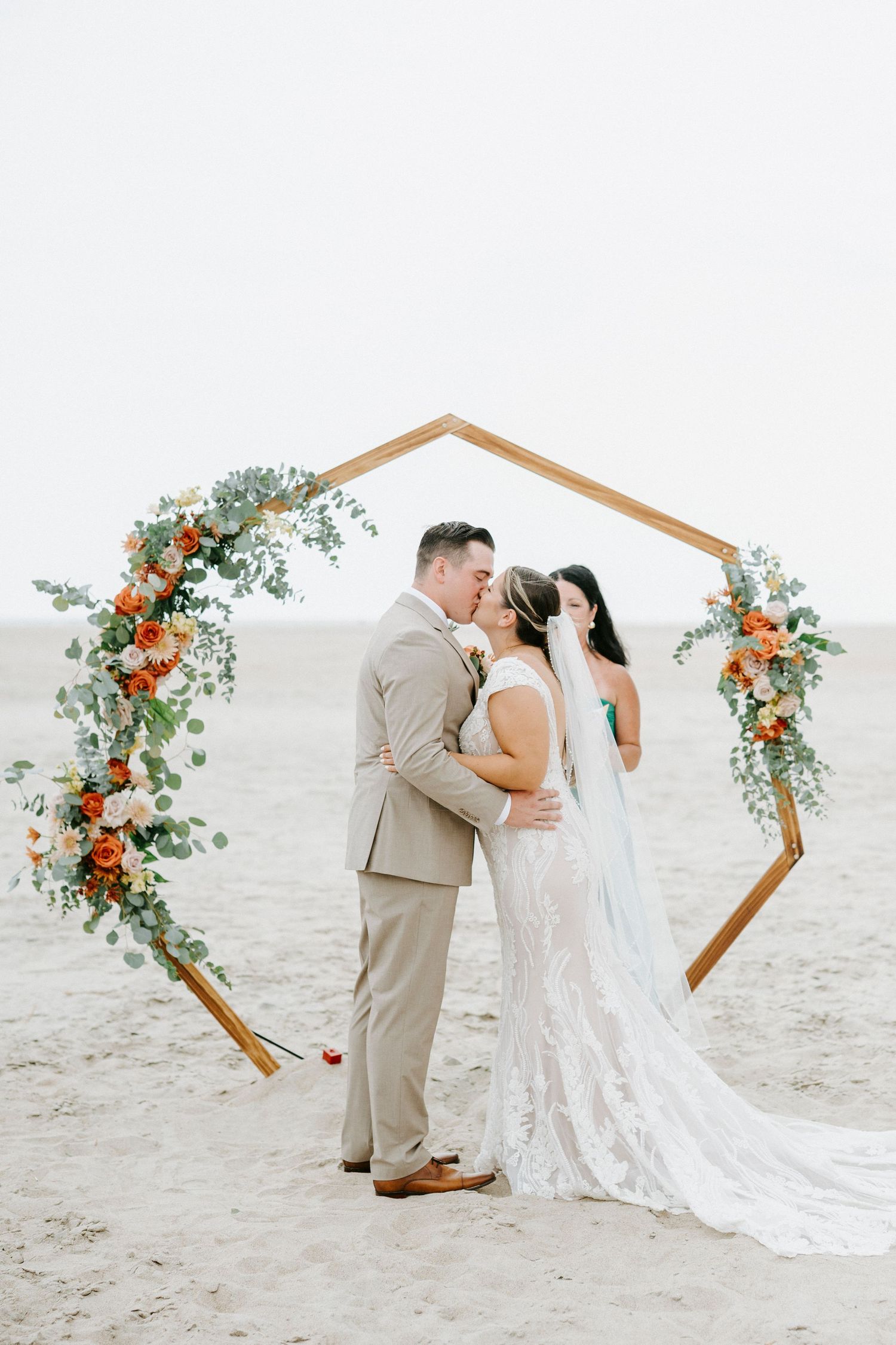 A beautiful beach wedding ceremony with a hexagonal wooden arch adorned with coral and peach florals on a sandy shoreline.