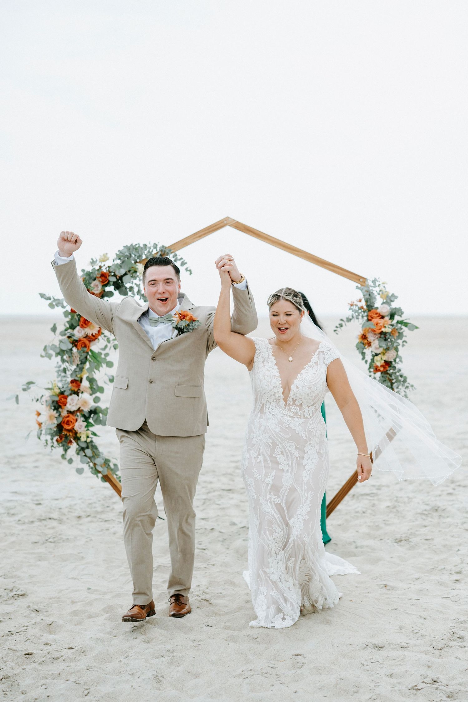 Newlywed couple and wedding party celebrate joyfully while walking on a sandy beach with coral flower arrangements.