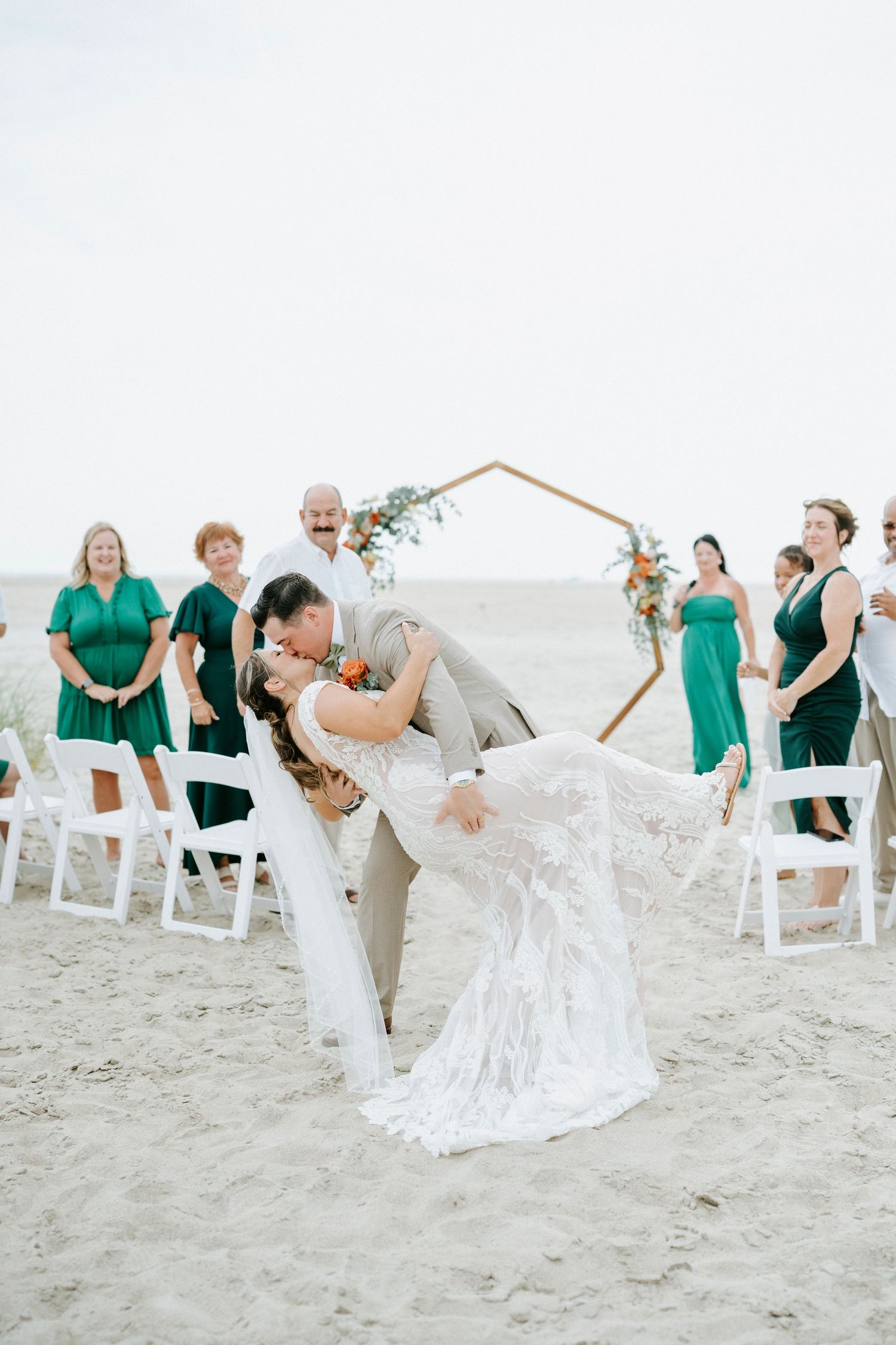Wedding guests in white and emerald green attire watch ceremony from wooden chairs on beach.