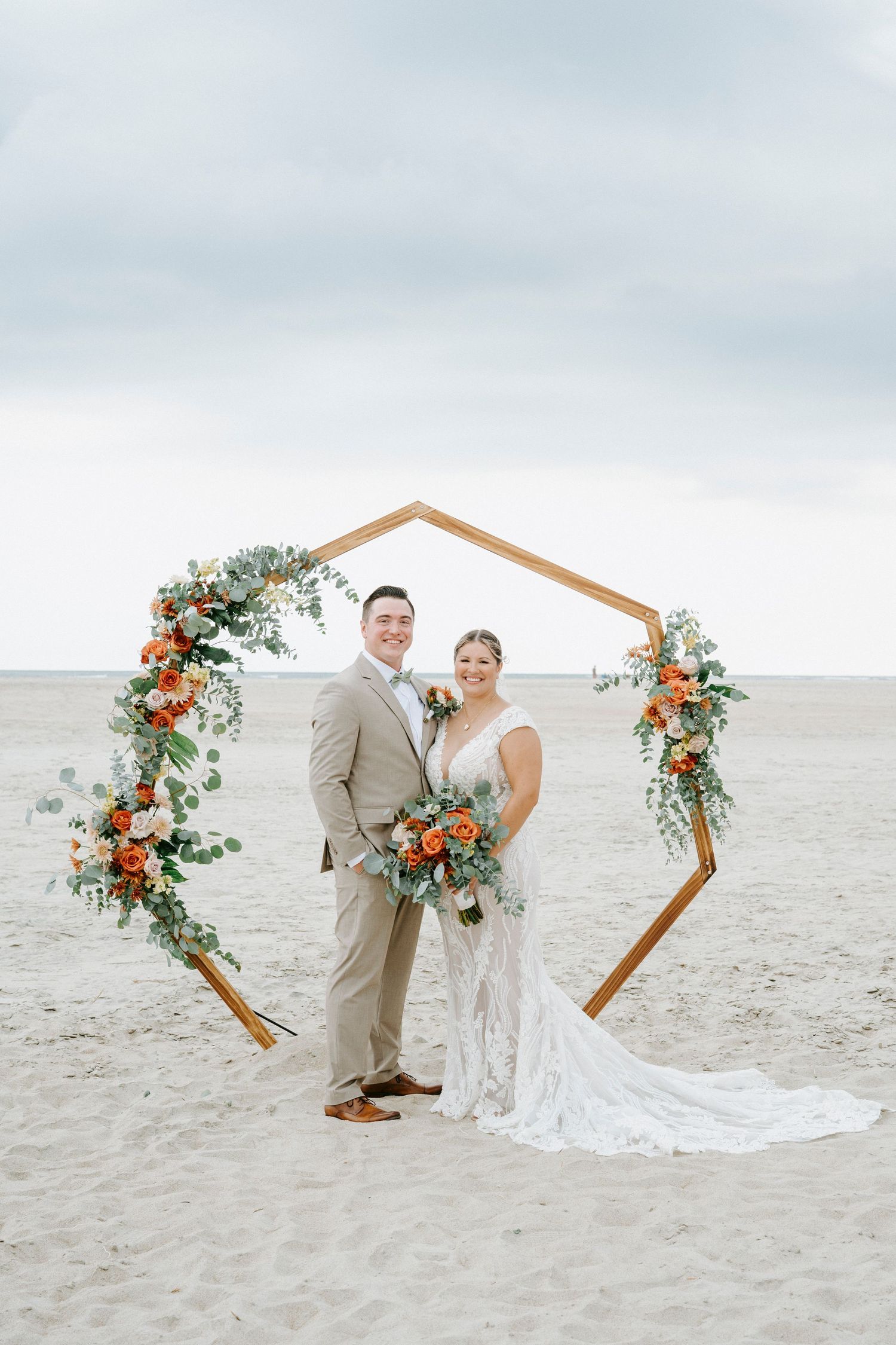 A beachside wedding ceremony setup with a hexagonal arch decorated with coral and white flowers on a sandy shore.