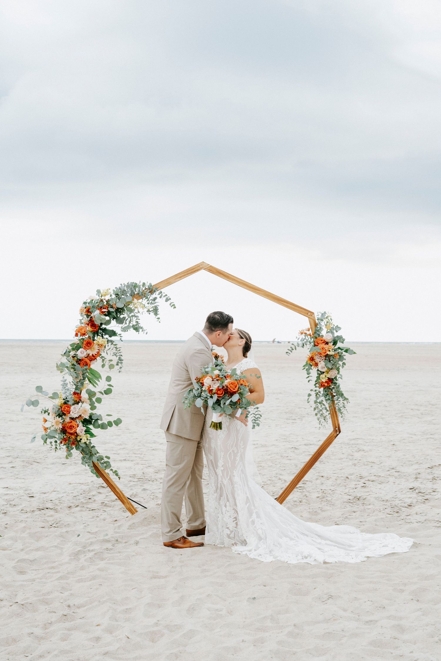 Couple sharing a romantic moment under a hexagonal floral arch on a serene beach during their wedding ceremony.