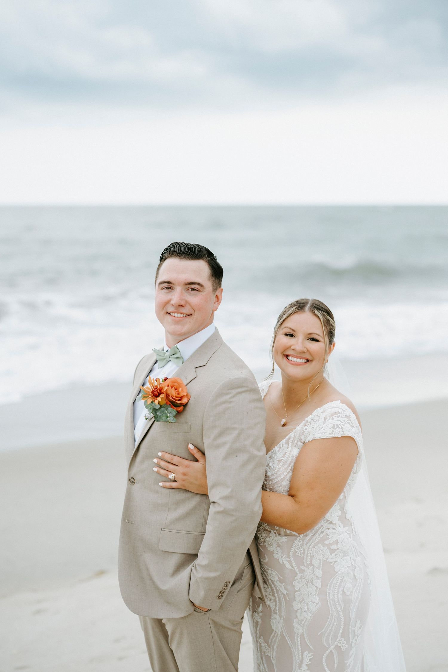 A beautiful beach wedding couple poses on the sand with ocean waves in the background on a cloudy day.