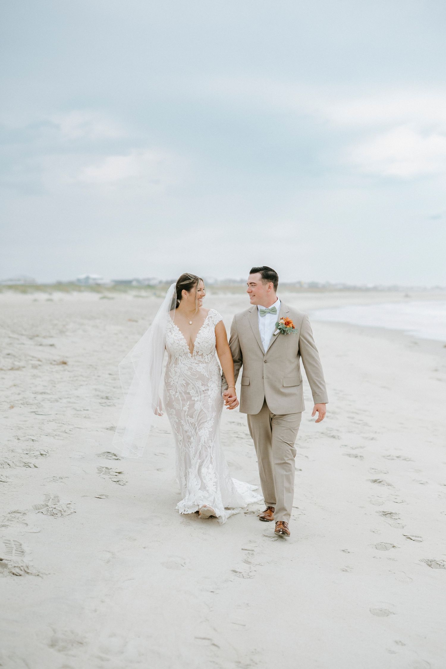 Newlyweds walking hand in hand along a pristine beach shoreline in elegant wedding attire.