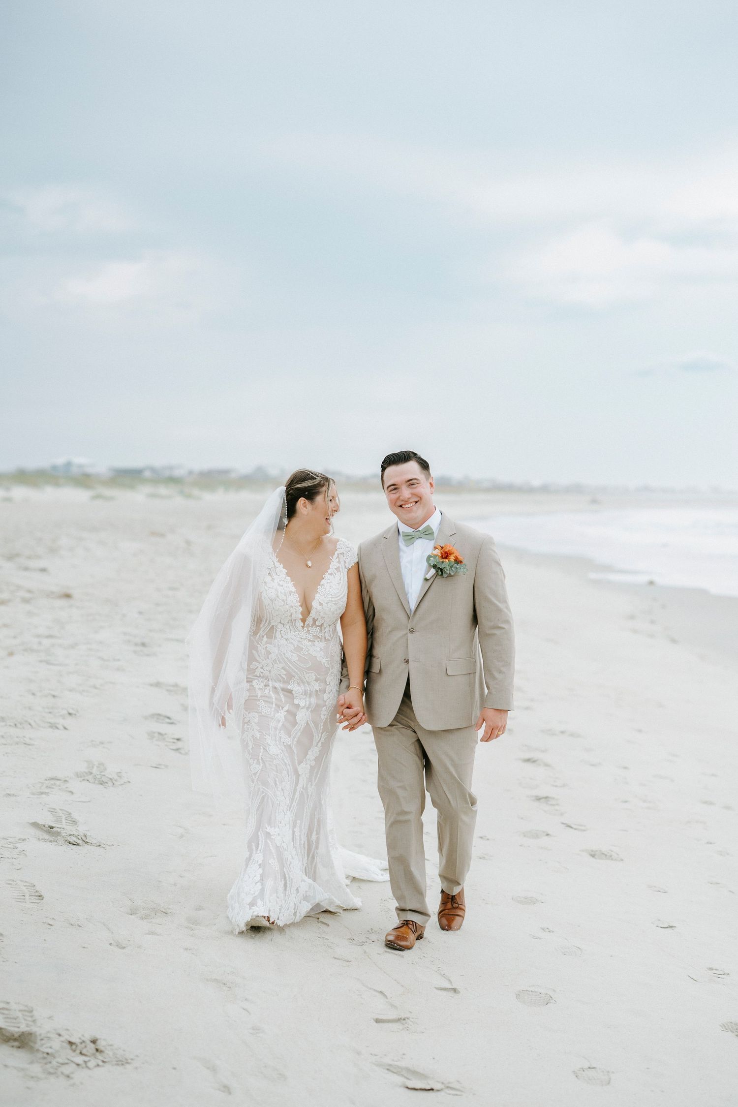 A romantic newlywed couple walks along a sandy beach shoreline in elegant wedding attire on an overcast day.