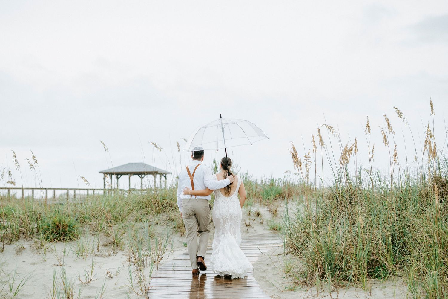 Wedding couple walking through beach grass and sand dunes with boardwalk in background.