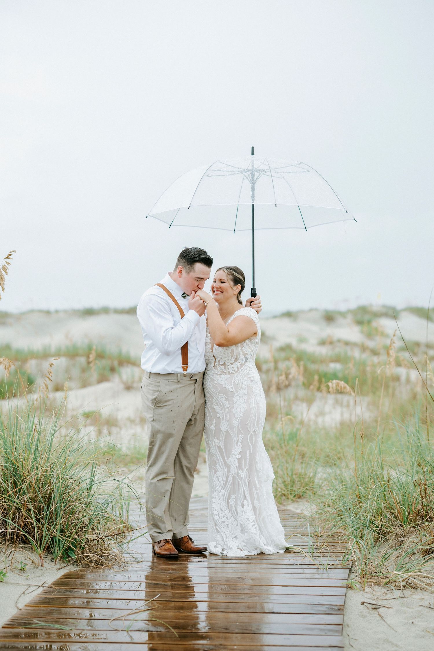 Romantic couple shares tender moment under clear umbrella on coastal boardwalk.