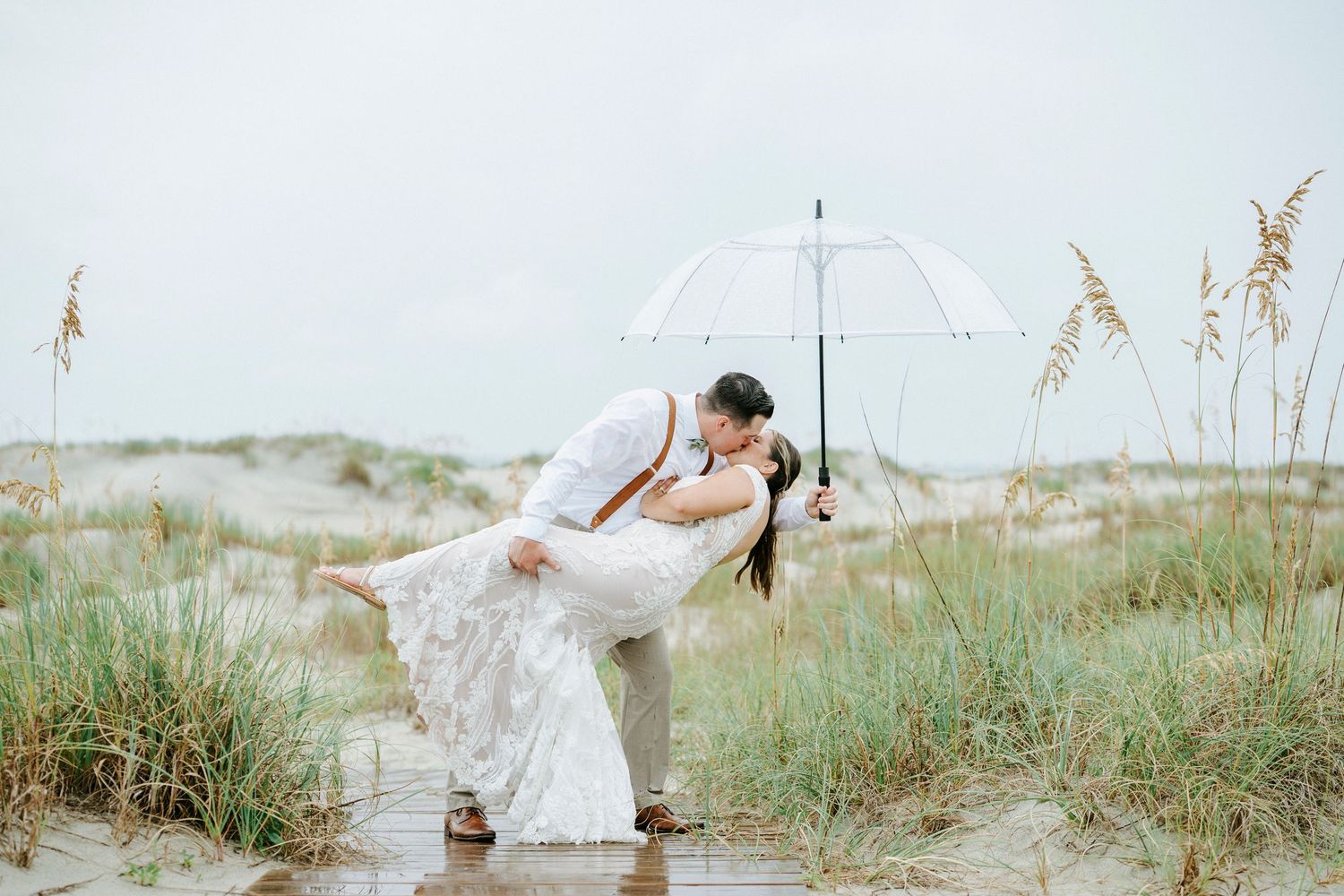 Clear umbrella frames wedding couple's embrace on wooden walkway through beach grass.