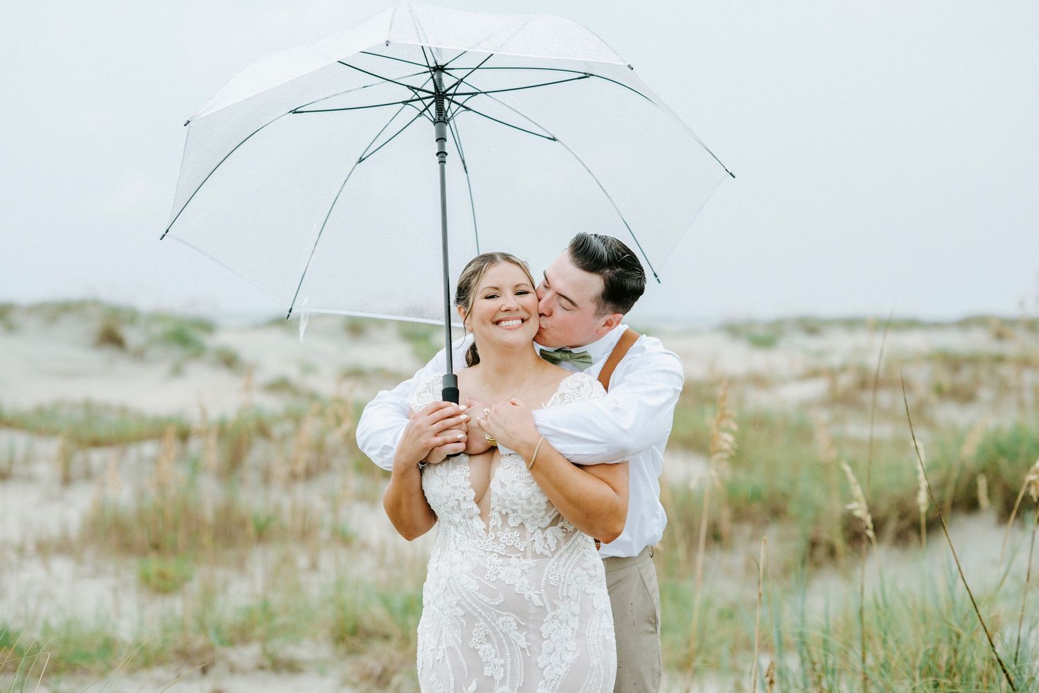 Clear umbrella shelters wedding couple's romantic moment on sandy coastal path.