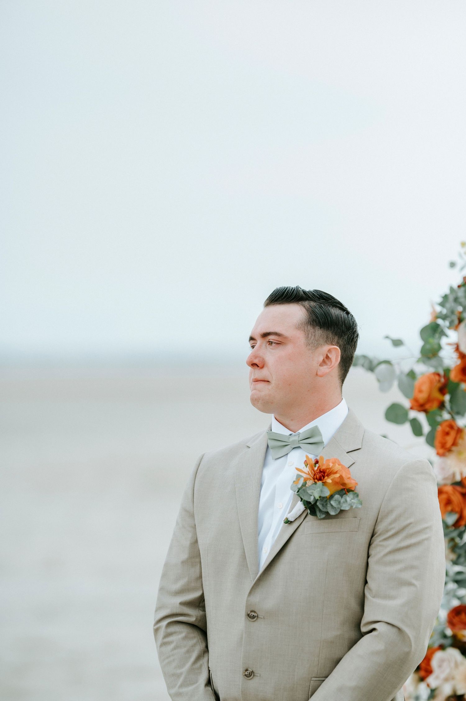 A groom in a light beige suit with peach boutonniere looks out pensively against an ocean backdrop.