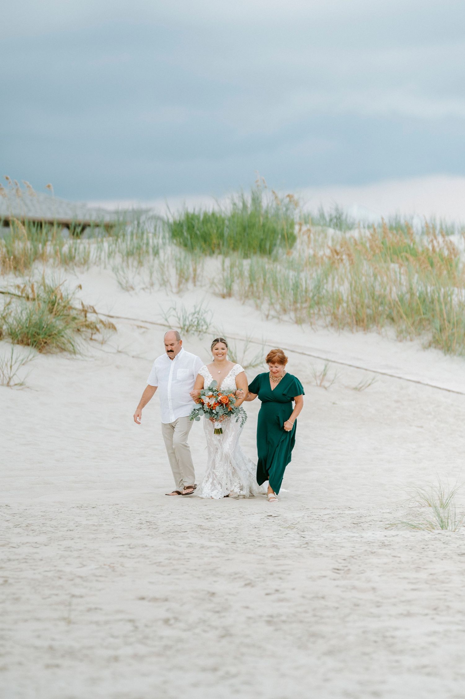 Wedding party walking together on a sandy beach path with sea grass and cloudy sky.