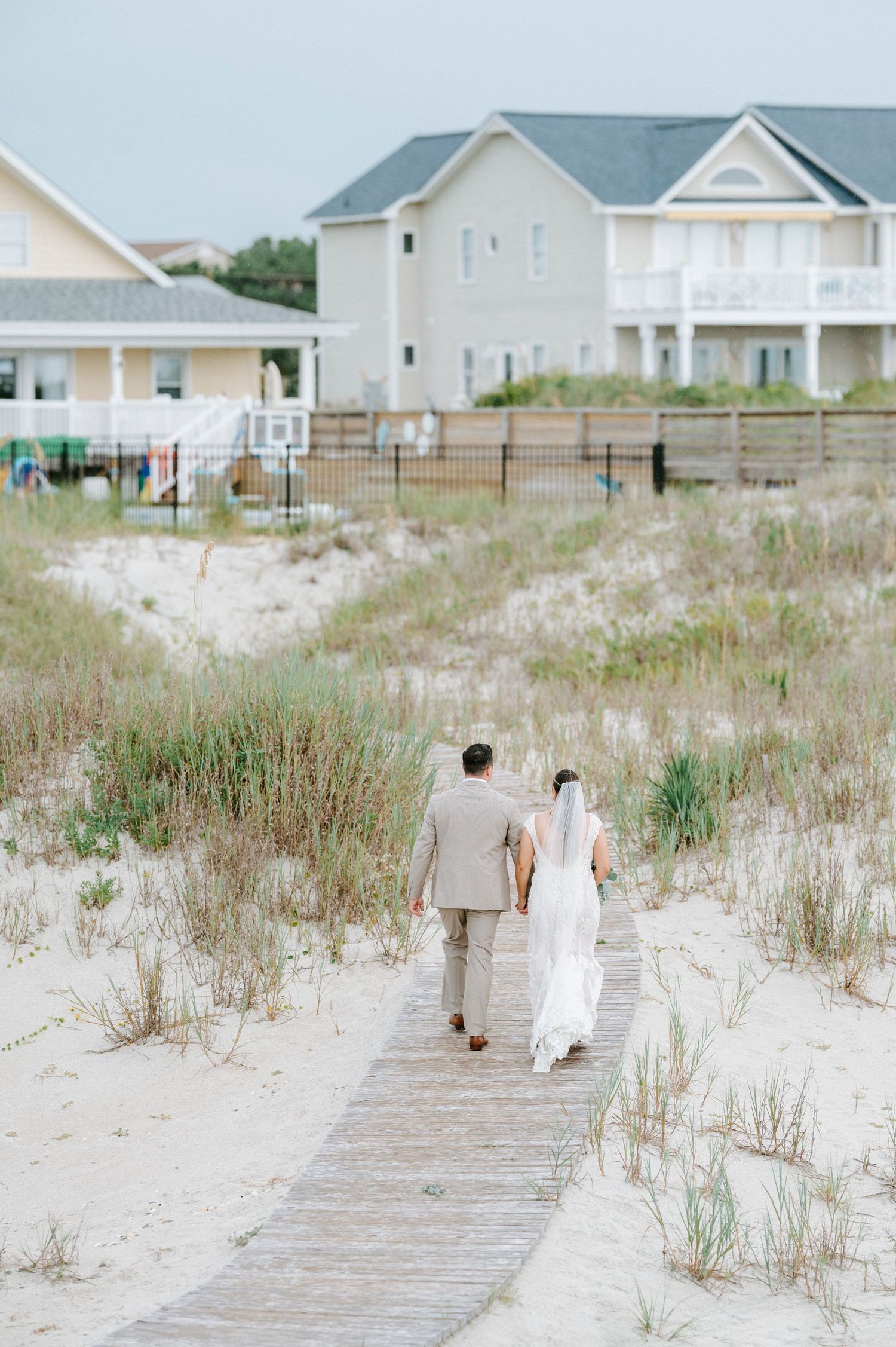 Coastal wedding party walks along sandy beach boardwalk with beach houses in the background.
