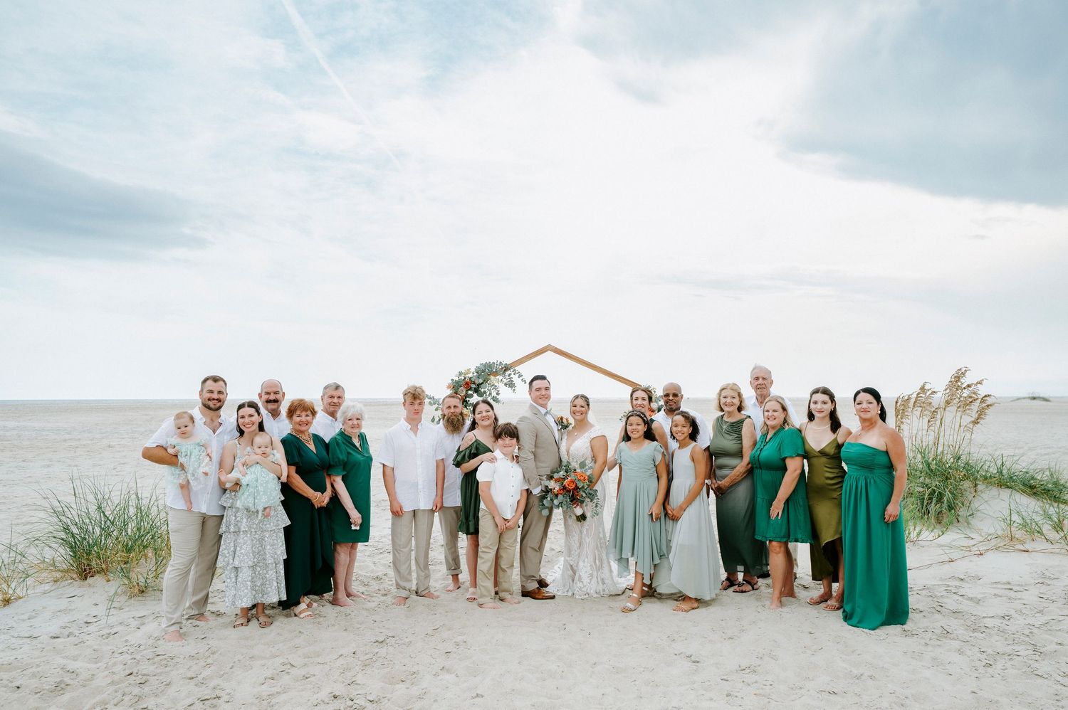 Wedding party group photo on a beach with bridesmaids in emerald green dresses and groomsmen in tan suits.