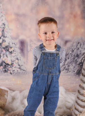 Young child wearing denim overalls and striped shirt poses in front of wintery backdrop with snow and sparkles.