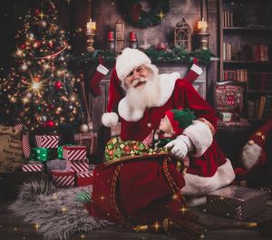 A person in a Santa Claus costume sits in a classic wooden sleigh surrounded by Christmas decorations and wrapped gifts.