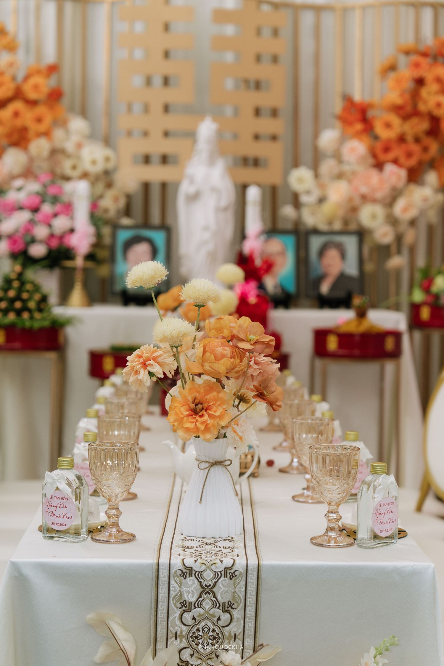 Elegant wedding table setting with orange ranunculus centerpiece, crystal glasses, and religious altar in background.