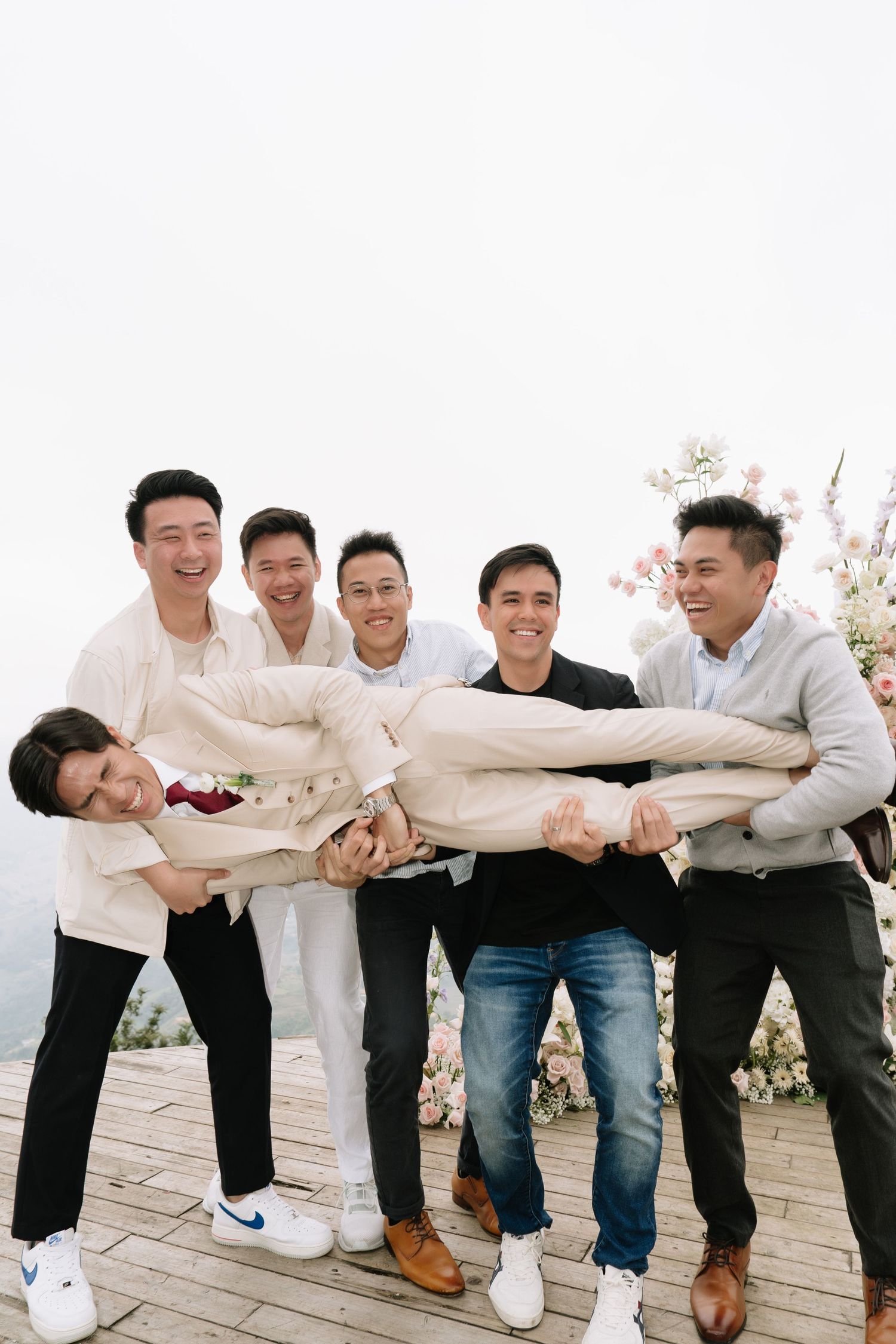 Groomsmen joyfully lifting groom horizontally at outdoor wedding venue with floral decorations in background.